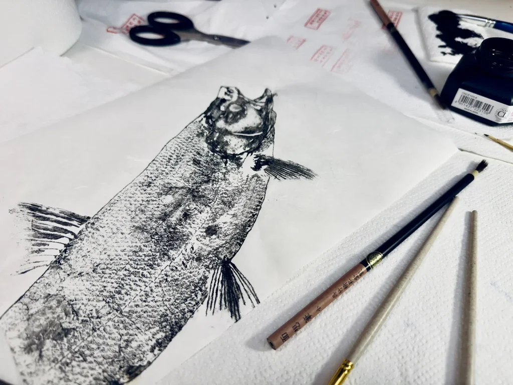 Odbitka ryby wykonana techniką gyotaku na papierze, obok pędzli i tuszu na stole roboczym.