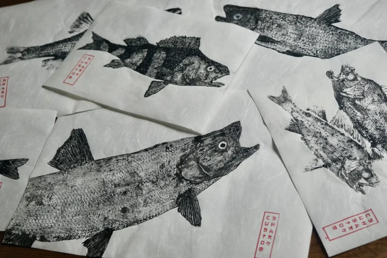 Odbitki gyotaku ryb o różnych kształtach i rozmiarach na zachodzących na siebie arkuszach papieru.